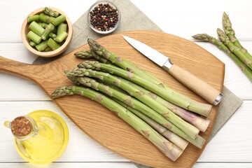 Fresh raw asparagus spears, knife and oil on white wooden table, flat lay