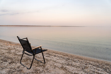 A single black folding chair faces the calm sea on an empty beach, captured at sunset with gentle...