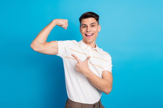 Young man in a white polo shirt flexing his arm and pointing on a blue background, expressing confidence and happiness
