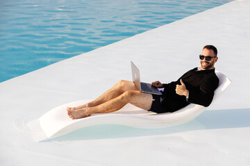 Man in blazer, swim trunks, and glasses sitting by the pool with a laptop, showing a thumbs-up...
