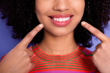 Smiling young woman gesturing with fingers to highlight her perfect teeth against vibrant purple...
