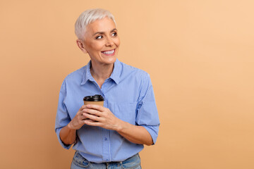 Smiling mature woman enjoys coffee in a casual blue striped shirt against a beige background in a...
