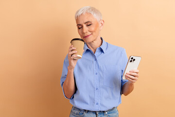 Mature woman with short white hair enjoying coffee while holding a smartphone against a beige...