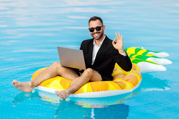 Man in a suit with a laptop sitting in a pool and showing an OK hand gesture. Concept confidence,...