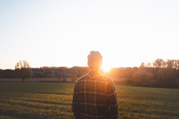 Young man in plaid shirt and beanie standing on green meadow at sunset. Golden sunlight, autumn...