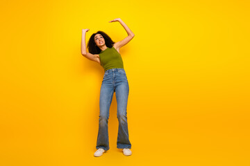 Young woman with curly hair in casual green top and jeans posing joyfully against a vibrant yellow...