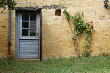 Porta in legno azzurro su parete rustica in pietra gialla, con rosa rossa rampicante e numero civico 1892, atmosfera romantica e rurale in un villaggio storico della campagna francese