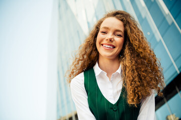 Confident young businesswoman in formal attire with curly red hair, smiling outdoors near a modern...