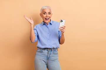 Happy senior woman gesturing excitedly while holding a smartphone against beige background