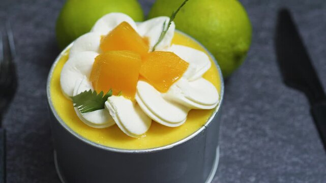 Delicate mango mousse cake Tin Can garnished with cream rosettes, fresh mango cubes, mint leaves, resting on dark gray textured surface near limes and kitchen knife