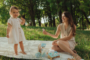 Happy young mother playing with her baby on the grass in a sunny park. Family bonding, early learning, parenting, love, outdoor lifestyle, childhood development, authentic real life moment.