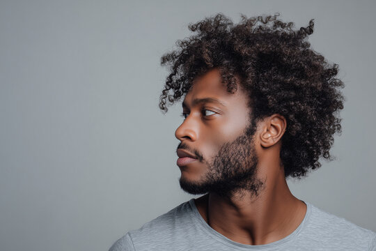 Profile portrait of a young man with curly hair and beard looking sideways against a neutral background - Powered by Adobe
