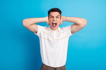 Frustrated young man wearing casual clothing covering ears with hands against vibrant blue...