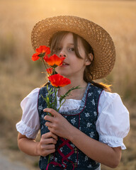Close up of adorable girl in dirndl and straw hat holding red poppies near her face. Traditional...