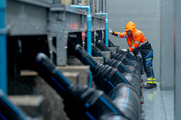 Wide shot of factory worker or technician with raincoat walk along pipe system and look up to maintenance in area of factory building during raining.
