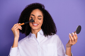 Smiling young woman applying makeup while holding compact mirror, posing in stylish attire on...