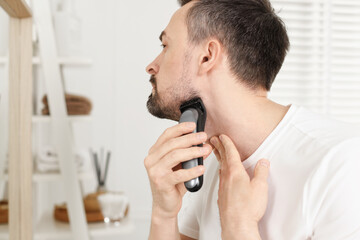 Handsome man shaving with trimmer near mirror at home