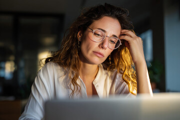 Thoughtful young woman with glasses working late on laptop in a dimly lit room, focused and concerned expression