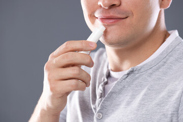 Young man applying lip balm onto his lips on grey background, closeup