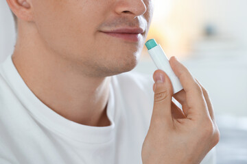 Young man applying lip balm onto his lips at home, closeup