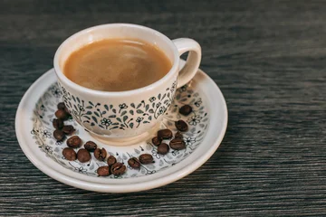 Fotobehang Koffiebonen Tasse en émail blanc avec des fleurs bleues remplie de café posée sur une soucoupe contenant des grains de café le tout sur une table grise  © Damien