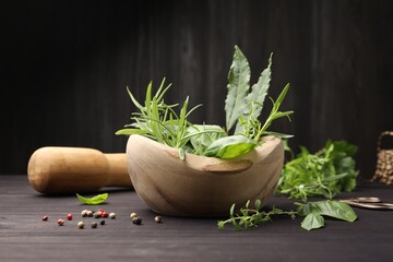 Mortar with different fresh herbs, peppercorns and pestle on wooden table against dark background,...