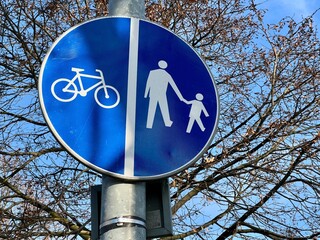 Blue traffic sign showing separated pedestrian and bicycle paths, close-up against tree branches and clear blue sky. Urban infrastructure, transportation, cycling, walking, outdoor signage.