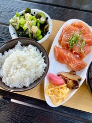 Vertical photo of traditional Japanese lunch set on dark wooden table with fresh sashimi, rice, miso soup, pickled vegetables, seaweed salad, and melon. Asian cuisine, healthy eating, authentic meal.