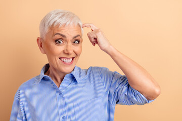 Confident Senior Woman Gesturing Towards Head Smiling in Blue Striped Shirt Against Beige Background