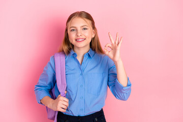 Happy schoolgirl with backpack making an OK gesture on pink background, showcasing a positive...