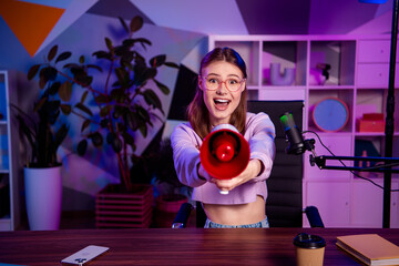 Young woman excitedly holding megaphone while recording podcast in trendy home studio with neon...