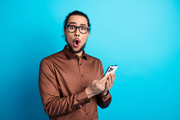 Young man in formalwear holding a smartphone with a surprised expression against a teal background