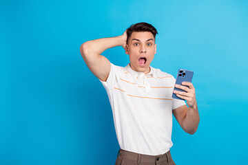 Surprised man holding smartphone with astonished expression against blue background in casual...