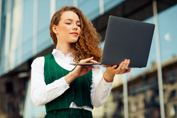 Cheerful businesswoman using a laptop outdoors near a modern building, showcasing professionalism