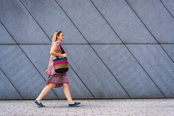 Beautiful mid adult woman walking along sidewalk in city against background of gray wall in summer....