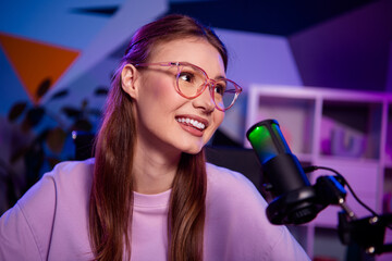 Confident young woman with glasses recording a podcast in a modern studio with dynamic colors and...
