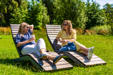 Two beautiful mature women sitting on park benches and talking in city park on sunny summer day
