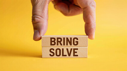 A person s fingers hold two wooden blocks stacked together with the words bring and solve carved into them against a bright yellow background