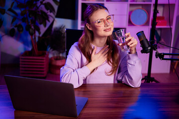Charming female blogger with a glass of water recording a podcast at home with a microphone and a...