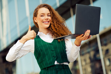 Confident businesswoman with curly red hair outdoors holding a laptop showing approval with a smile...