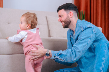 Loving father kneels on carpet supporting daughter in pink overalls as she practices standing up,...