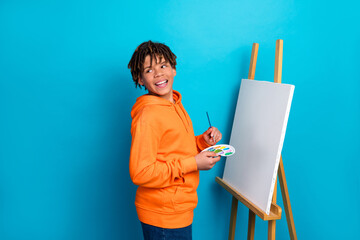 Teen in bright hoodie creates art on canvas while standing against vivid blue background,...