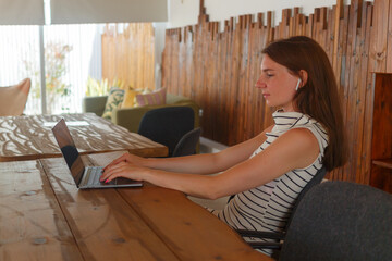 A young woman is focused on her laptop in a cozy wooden cafe, surrounded by natural light and...