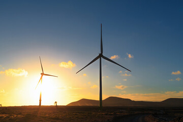 Two wind turbines stand tall against a vibrant sunset, with mountains and clouds in the background...