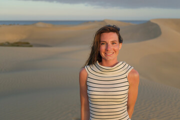 A woman stands confidently in a vast desert, smiling as the sun sets, casting warm colors over the...