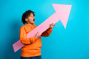 Delighted boy in orange hoodie holds a pink arrow prop, showcasing enthusiasm against a bright blue...