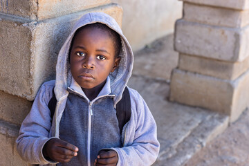 Africa village, single african child in school uniform with hoodie, sited in the yard in front of...