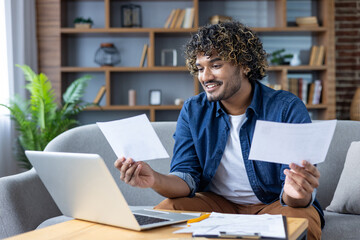 A young man reviews documents while using his laptop in a comfortable home setting, smiling and...
