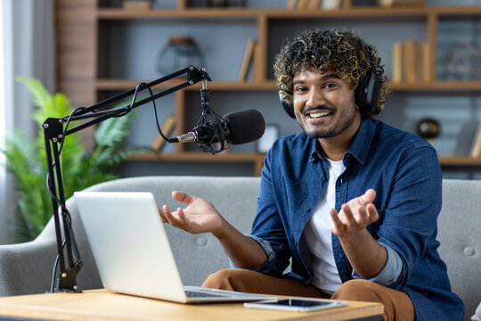 A smiling man with curly hair records a podcast at home, using a microphone and laptop.