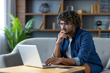 A young man with curly hair and glasses works on a laptop, deep in thought, in a modern living room.